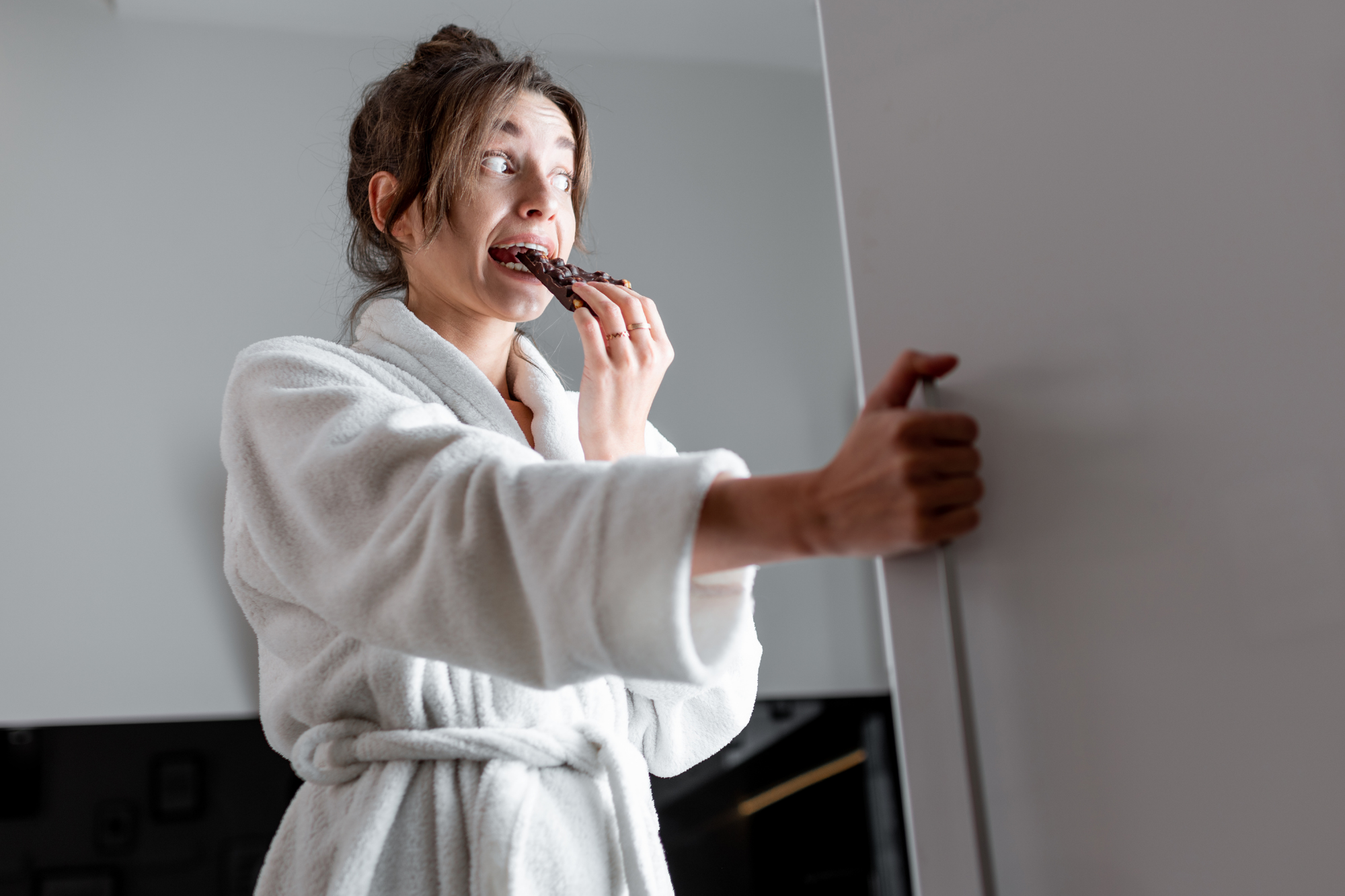 girl looking in fridge holding chocolate wondering how to stop overeating