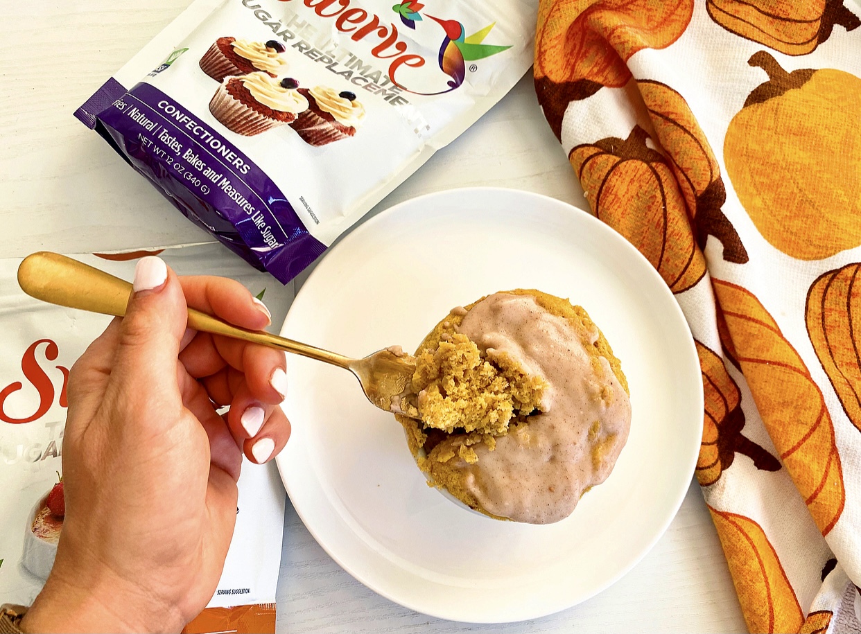 Hand holding a fork taking a piece of low carb pumpkin spice mug cake. Pumpkin mug cake is on a white plate next to a pumpkin napkin and Swerve confectioners sugar and granulated sugar bags. 